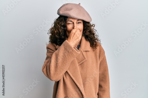 Middle age hispanic woman wearing french look with beret smelling something stinky and disgusting, intolerable smell, holding breath with fingers on nose. bad smell
