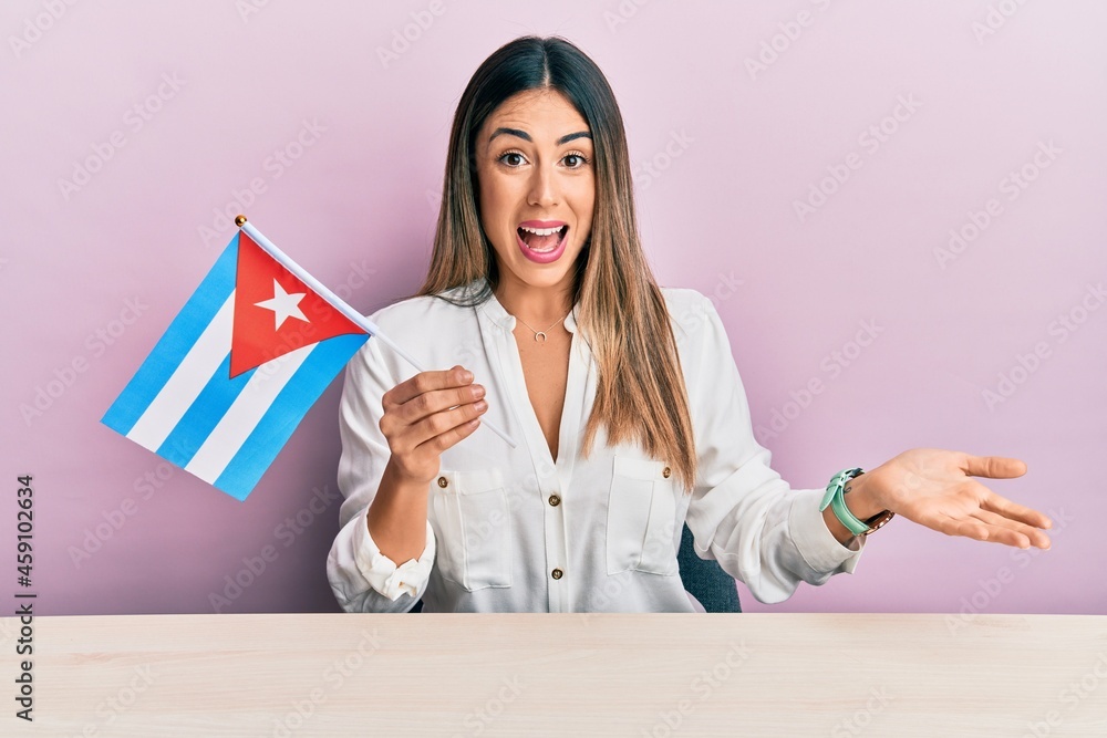 Young hispanic woman holding cuba flag sitting on the table celebrating achievement with happy smile and winner expression with raised hand Young hispanic woman holding cuba flag sitting on the table celebrating achievement with happy smile and winner expression with raised hand
