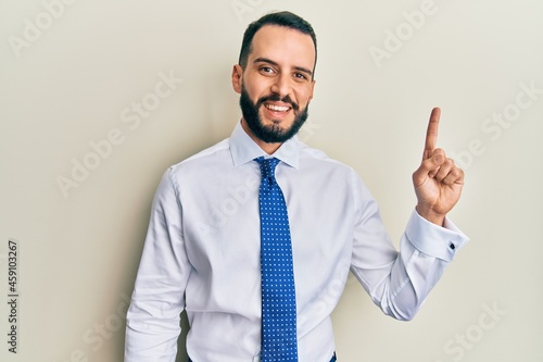 Young man with beard wearing business tie showing and pointing up with finger number one while smiling confident and happy.