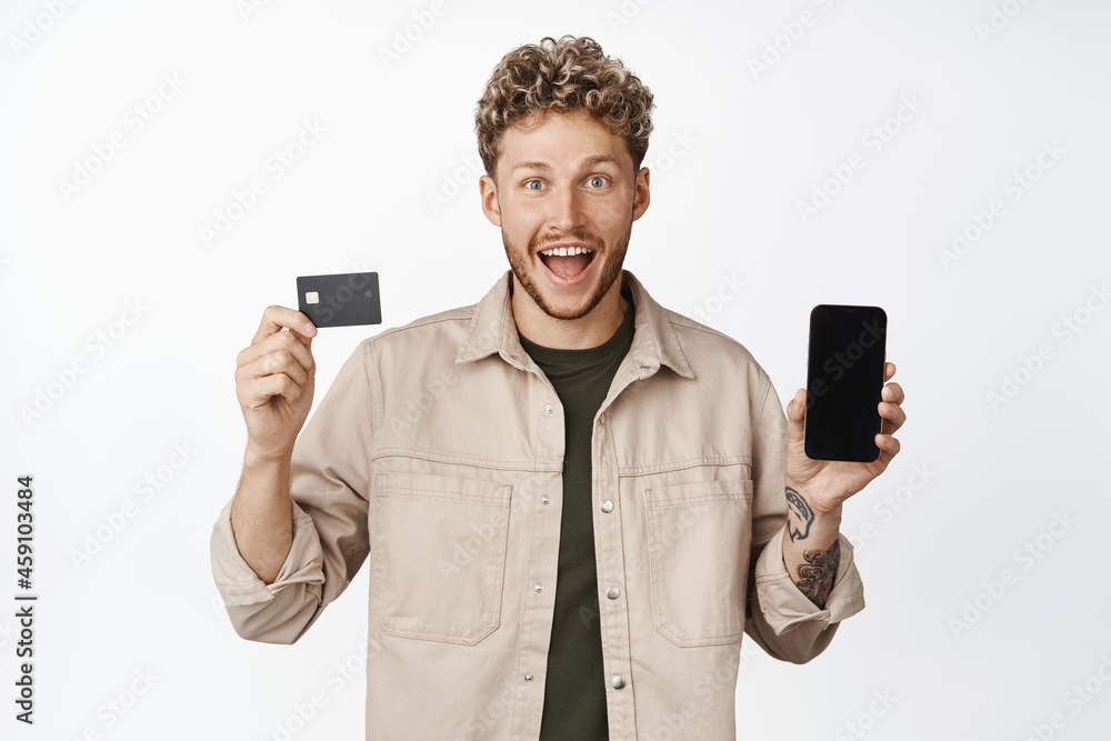Young happy blond man showing credit card with mobile phone screen, smiling amazed, announcing awesome promotion, standing over white background Young happy blond man showing credit card with mobile phone screen, smiling amazed, announcing awesome promotion, standing over white background