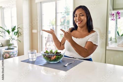 Young hispanic woman eating healthy salad at home amazed and smiling to the camera while presenting with hand and pointing with finger.