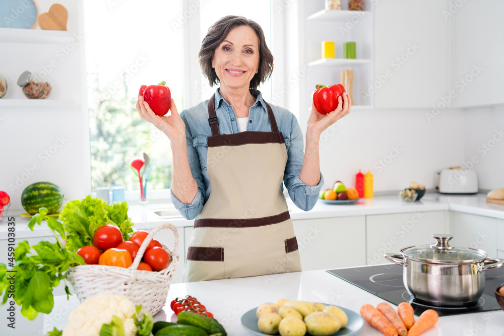 Photo of cheerful positive charming old woman hold hands peppers cook food indoors inside house home kitchen