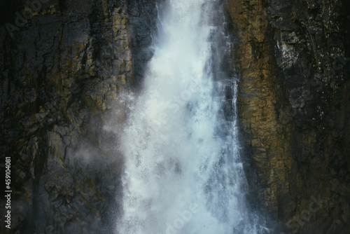 Atmospheric minimal landscape with vertical big waterfall on rock mountain wall. Powerful large waterfall in dark gorge. Nature background of high vertical turbulent falling water stream on wet rocks.