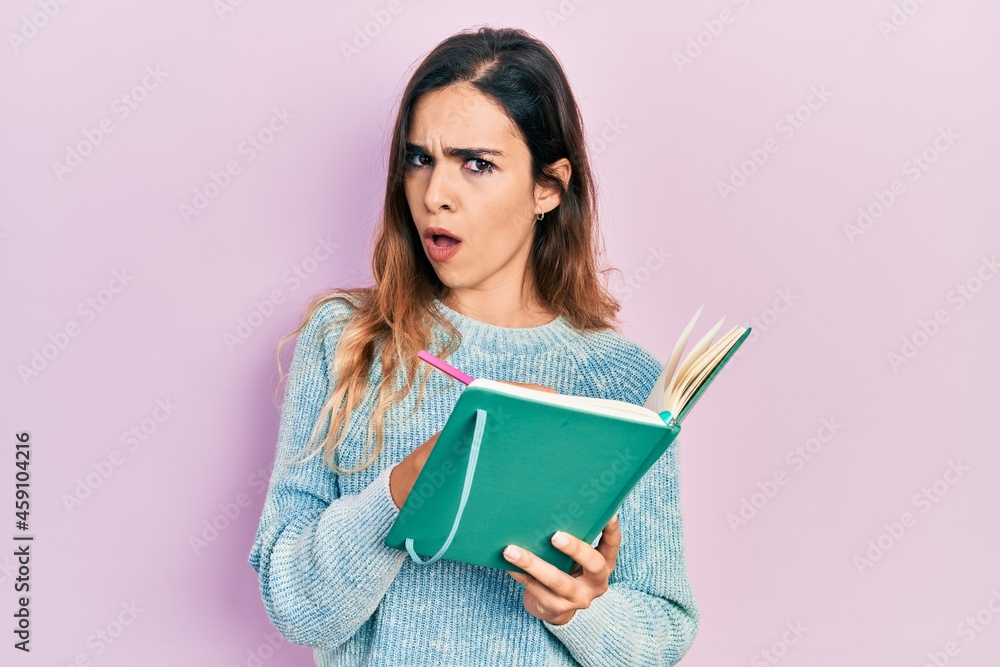Young hispanic girl reading and writing book in shock face, looking skeptical and sarcastic, surprised with open mouth Young hispanic girl reading and writing book in shock face, looking skeptical and sarcastic, surprised with open mouth