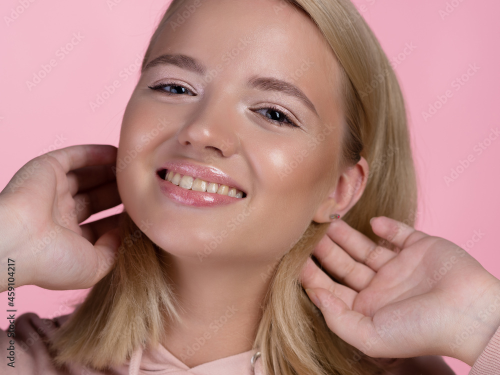 cute young blonde in a pink hoodie smiles happily and touches her hair with her hand. cute young blonde in a pink hoodie smiles happily and touches her hair with her hand.