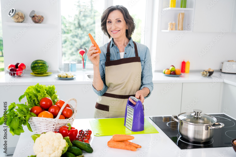 Photo of age cheerful lady cut carrot wear apron jeans shirt at kitchen alone Photo of age cheerful lady cut carrot wear apron jeans shirt at kitchen alone