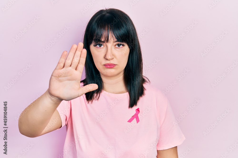 Young hispanic woman wearing pink cancer ribbon on shirt with open hand doing stop sign with serious and confident expression, defense gesture