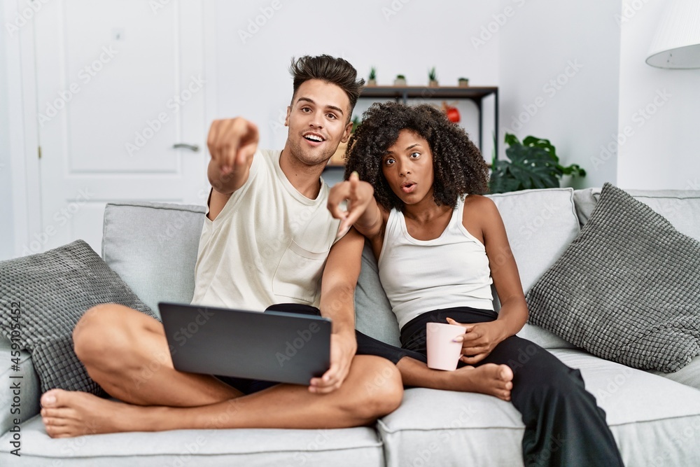 Young interracial couple using laptop at home sitting on the sofa pointing with finger surprised ahead, open mouth amazed expression, something on the front Young interracial couple using laptop at home sitting on the sofa pointing with finger surprised ahead, open mouth amazed expression, something on the front