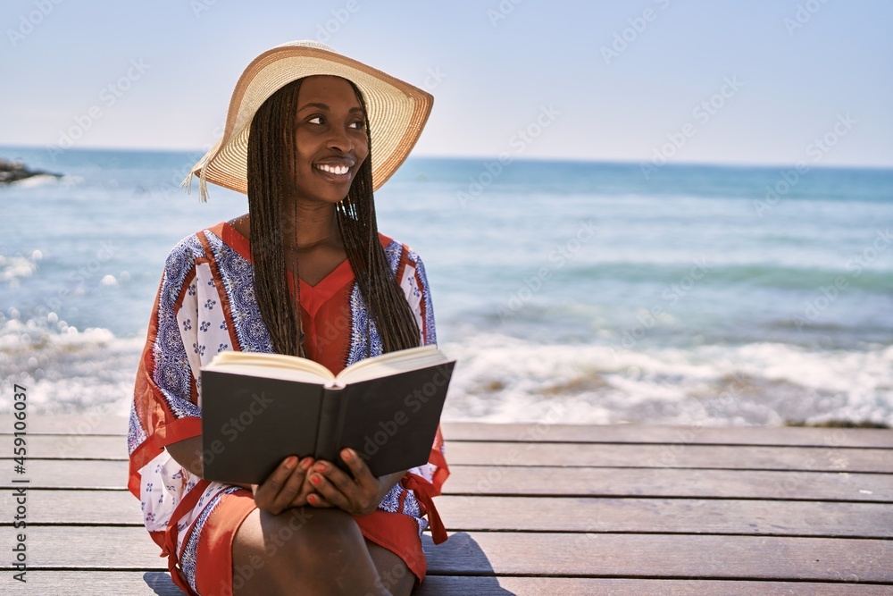 Young african american woman reading book sitting on the bench at the beach.