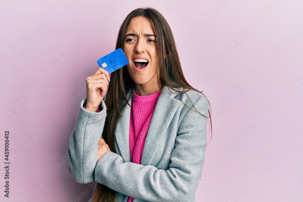 Young hispanic girl holding credit card angry and mad screaming frustrated and furious, shouting with anger. rage and aggressive concept.