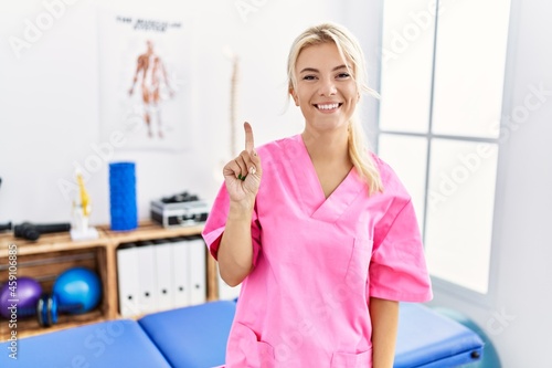 Young caucasian woman working at pain recovery clinic showing and pointing up with finger number one while smiling confident and happy.