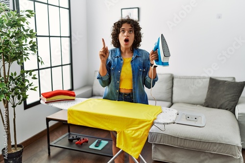 Young hispanic woman ironing clothes at home amazed and surprised looking up and pointing with fingers and raised arms.