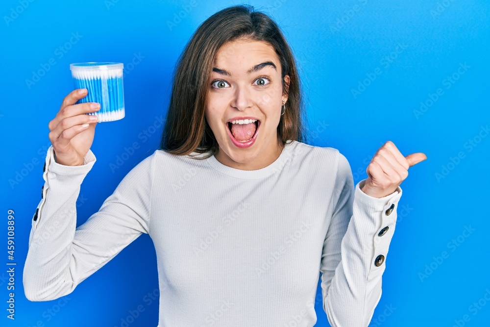 Young caucasian girl holding earwax cotton remover pointing thumb up to the side smiling happy with open mouth