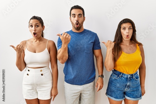 Group of young hispanic people standing over isolated background surprised pointing with hand finger to the side  open mouth amazed expression.