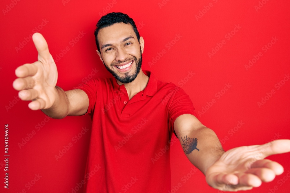 Hispanic man with beard wearing casual red t shirt looking at the camera smiling with open arms for hug. cheerful expression embracing happiness. Hispanic man with beard wearing casual red t shirt looking at the camera smiling with open arms for hug. cheerful expression embracing happiness.