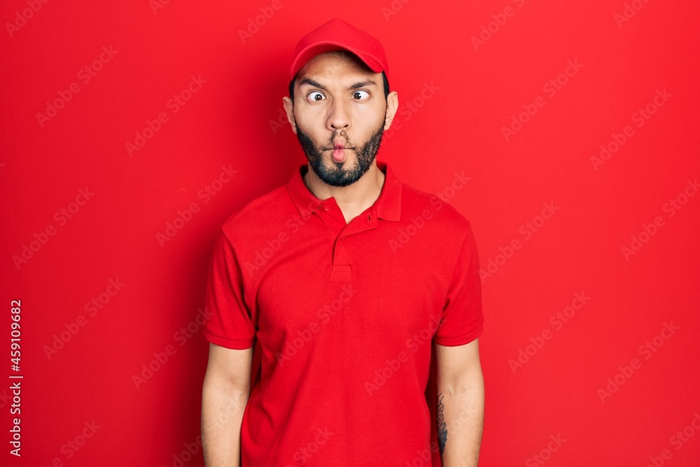 Hispanic man with beard wearing delivery uniform and cap making fish face with lips, crazy and comical gesture. funny expression. Hispanic man with beard wearing delivery uniform and cap making fish face with lips, crazy and comical gesture. funny expression.