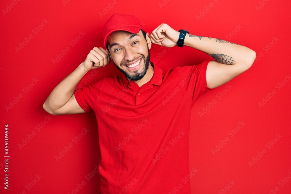 Fototapeta premium Hispanic man with beard wearing delivery uniform and cap smiling pulling ears with fingers, funny gesture. audition problem
