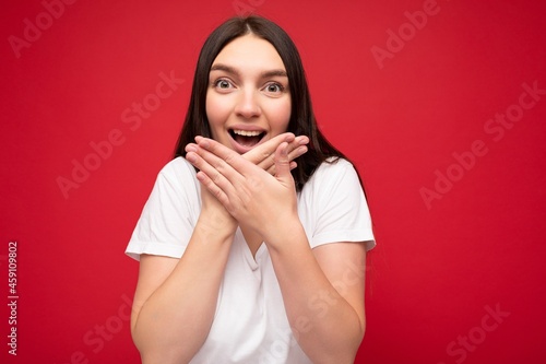 Photo of positive young beautiful brunette female person with sincere emotions wearing casual white t-shirt for mockup isolated over red background with copy space and covering mouth
