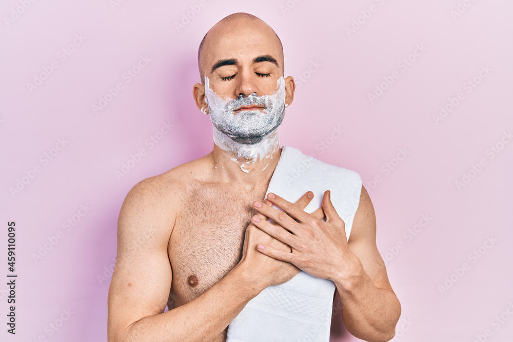 Young bald man shirtless shaving beard with foam smiling with hands on chest with closed eyes and grateful gesture on face. health concept.