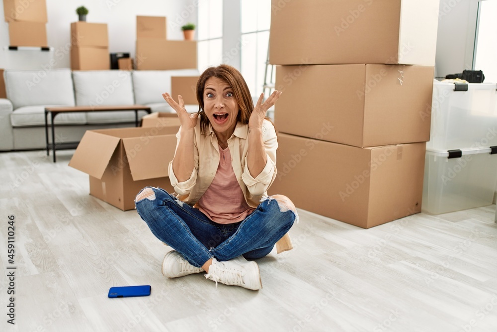 Senior middle east woman sitting on the floor at new home celebrating victory with happy smile and winner expression with raised hands Senior middle east woman sitting on the floor at new home celebrating victory with happy smile and winner expression with raised hands