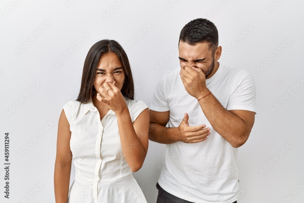 Young interracial couple standing together in love over isolated background smelling something stinky and disgusting, intolerable smell, holding breath with fingers on nose. bad smell Young interracial couple standing together in love over isolated background smelling something stinky and disgusting, intolerable smell, holding breath with fingers on nose. bad smell