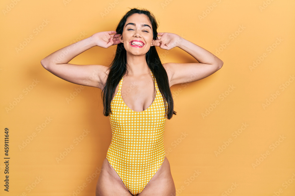 Young hispanic girl wearing swimsuit smiling pulling ears with fingers, funny gesture. audition problem Young hispanic girl wearing swimsuit smiling pulling ears with fingers, funny gesture. audition problem