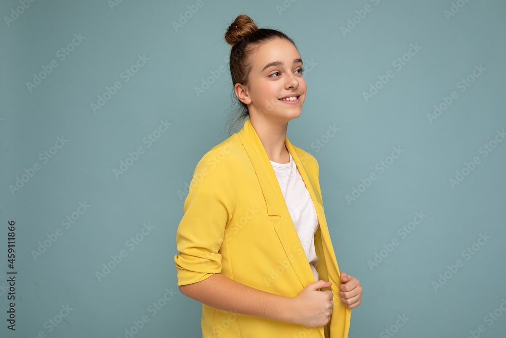 Side-profile shot of attractive positive happy smiling brunette little girl wearing casual yellow jacket and white t-shirt standing isolated over blue background wall looking to the side Side-profile shot of attractive positive happy smiling brunette little girl wearing casual yellow jacket and white t-shirt standing isolated over blue background wall looking to the side