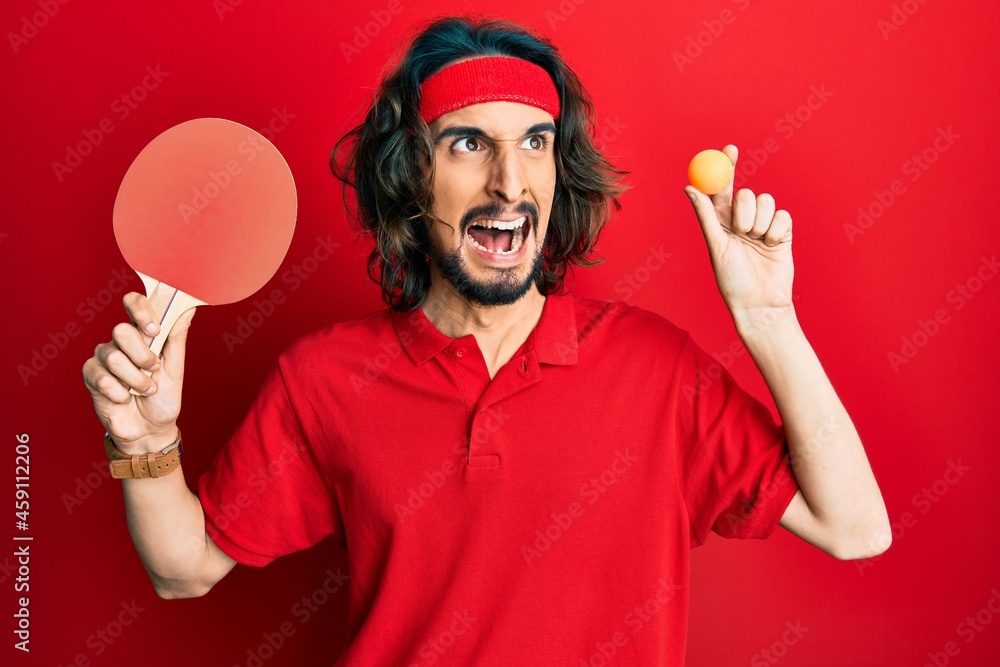 Young hispanic man holding red ping pong racket and ball angry and mad screaming frustrated and furious, shouting with anger. rage and aggressive concept. Young hispanic man holding red ping pong racket and ball angry and mad screaming frustrated and furious, shouting with anger. rage and aggressive concept.