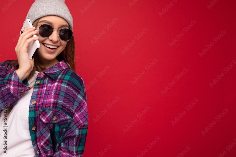 Closeup photo of beautiful happy positive young blonde woman wearing hipster purple shirt and casual white t-shirt grey hat and sunglasses isolated over red background holding in hand and talking on Closeup photo of beautiful happy positive young blonde woman wearing hipster purple shirt and casual white t-shirt grey hat and sunglasses isolated over red background holding in hand and talking on