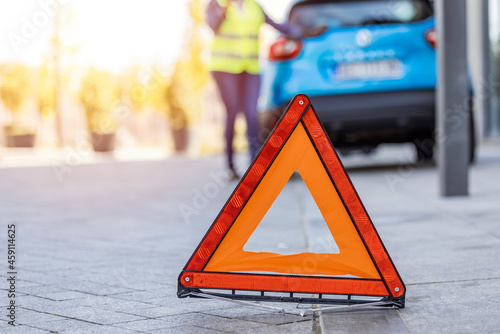 Close up on triangle warning sign with woman calling for assistance after breaking down with her car on background. Red triangle sign on the road and Young woman. Woman having car problems