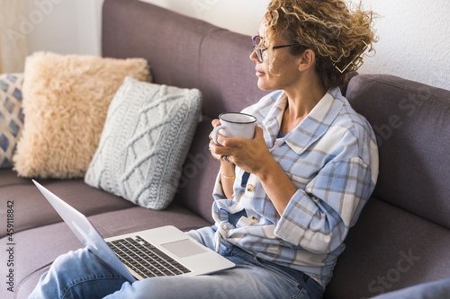 Beautiful young woman in eyeglasses working on laptop while drinking coffee sitting on sofa. Businesswoman in casuals looking away while working from home. Woman having coffee break while using laptop