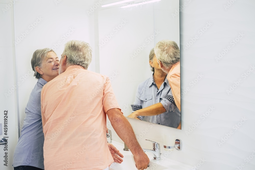 Obraz premium Romantic senior couple romancing while washing hands in bathroom at home. Elderly couple using bathroom. Loving old couple kissing each other in front of mirror in bathroom