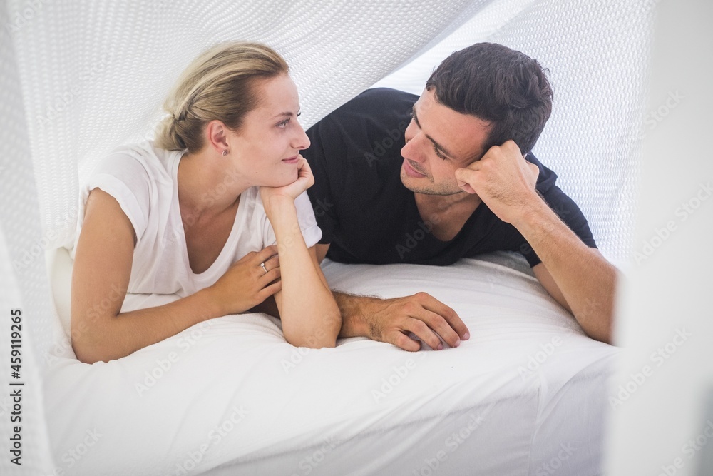 Young couple lying on front and staring at each other. Beautiful couple spending leisure with each other. Loving couple looking at each other on cozy bed at home