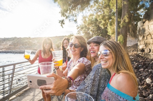 Group of female friends taking selfie on smartphone while holding glass of juice by sea. Young women enjoying summer holidays. Happy female friends enjoying drinks and taking selfie using mobile phone