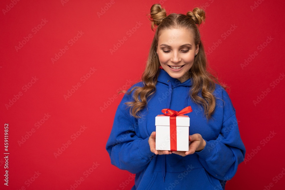 Shot of attractive happy smiling young blonde woman isolated over red background wall wearing blue trendy hoodie holding gift box and looking at white box with red ribbon