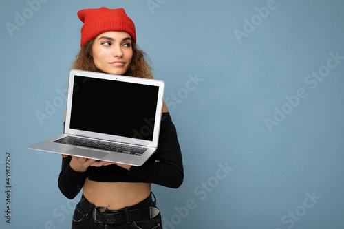 Side profile photo shot of beautifyl funny happy young brunet curly female teenager wearing black crop top and red and orange do-rag isolated over light blue wall background holding computer laptop photo