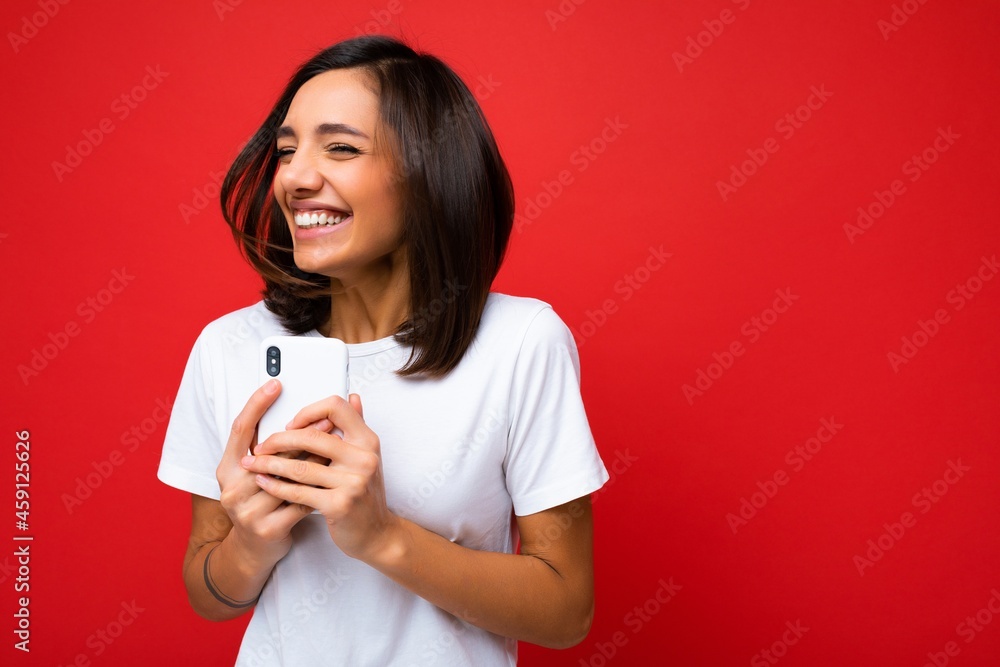 Photo shot of attractive positive happy smiling good looking young brunette woman wearing casual stylishwhite t-shirt poising isolated on red background with empty space holding in hand and using Photo shot of attractive positive happy smiling good looking young brunette woman wearing casual stylishwhite t-shirt poising isolated on red background with empty space holding in hand and using