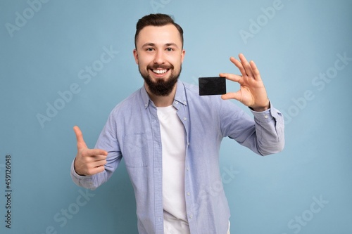 photo shot of Handsome smiling brunette bearded young man wearing stylish blue shirt and white t-shirt isolated over blue background wall holding credit card looking at camera
