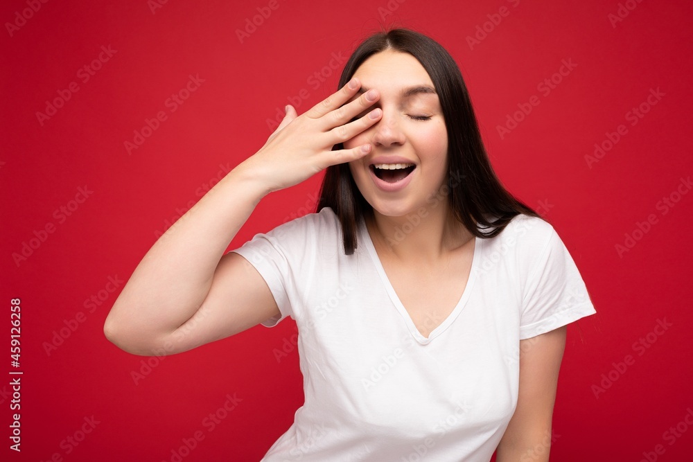 Photo of positive young beautiful brunette female person with sincere emotions wearing casual white t-shirt for mockup isolated over red background with copy space and covering eye Photo of positive young beautiful brunette female person with sincere emotions wearing casual white t-shirt for mockup isolated over red background with copy space and covering eye