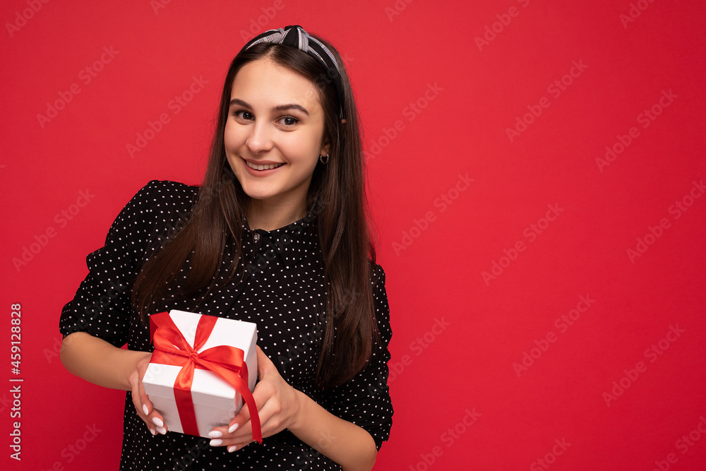 Shot of charming happy smiling brunette girl isolated over red background wall wearing black blouse holding white gift box with red ribbon and looking at camera Shot of charming happy smiling brunette girl isolated over red background wall wearing black blouse holding white gift box with red ribbon and looking at camera