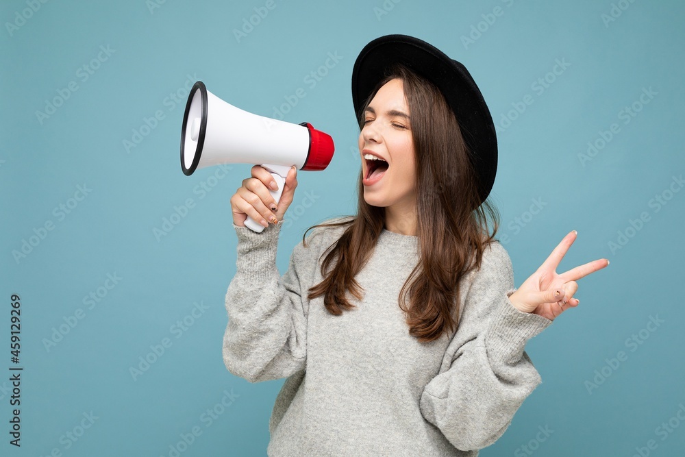 Photo of young beautiful positive brunette woman with sincere emotions wearing stylish black hat and grey sweater isolated over blue background with copy space and screaming in magaphone, shows peace Photo of young beautiful positive brunette woman with sincere emotions wearing stylish black hat and grey sweater isolated over blue background with copy space and screaming in magaphone, shows peace