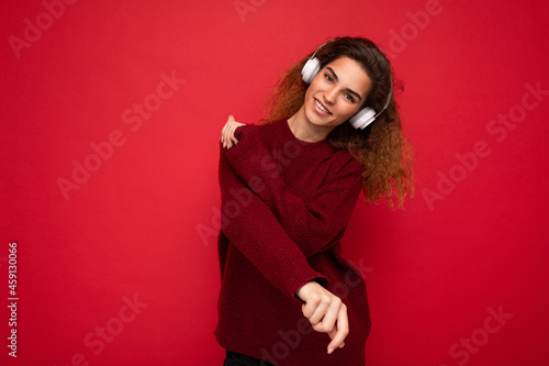 Beautiful happy smiling young brunette curly woman wearing dark red sweater isolated over red background wall wearing white bluetooth headphones listening to music and having fun looking at camera