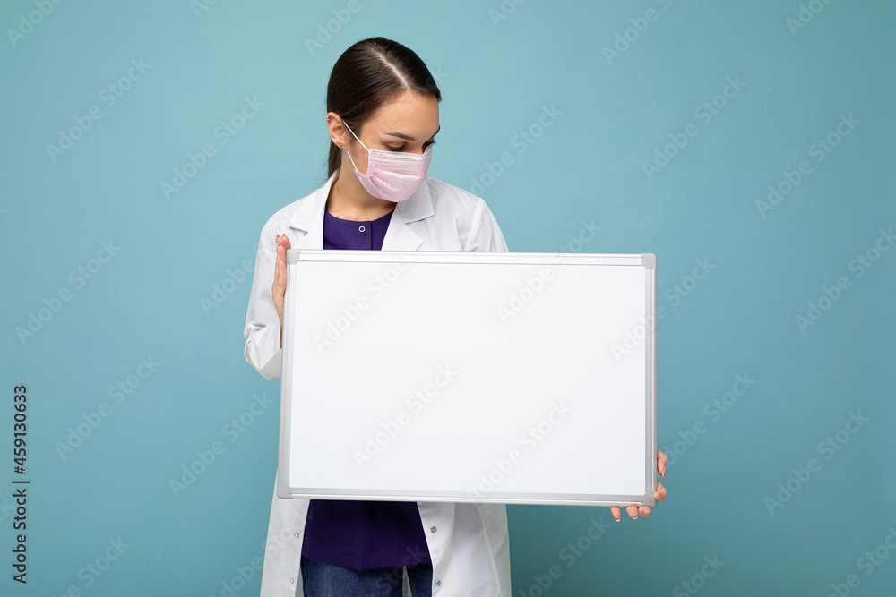 Woman doctor wearing a white medical coat and a mask holding blank board with copy space for text isolated on background. Coronavirus concept