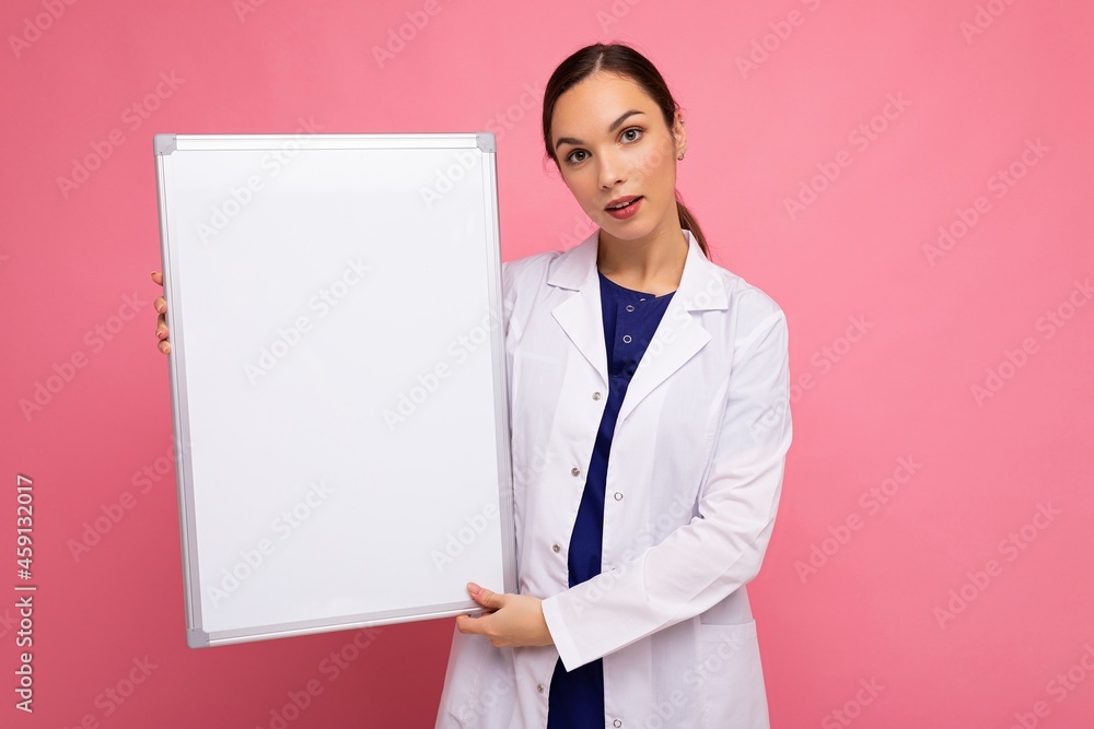 Portrait of attractive pretty young positive brunette woman wearing medical white coat holding white magnetic board for mock up isolated over pink background with copy space Portrait of attractive pretty young positive brunette woman wearing medical white coat holding white magnetic board for mock up isolated over pink background with copy space