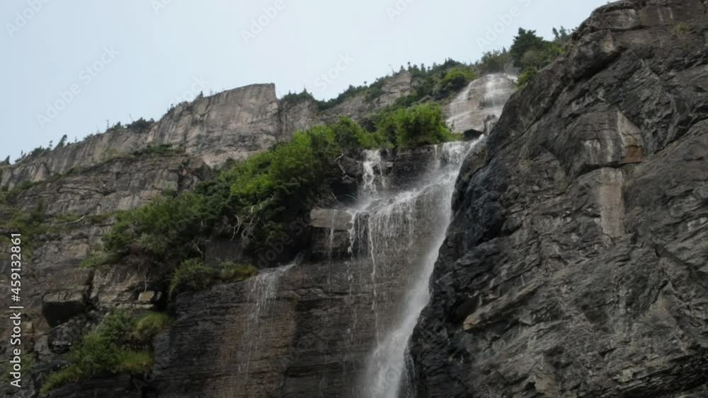 A waterfall rushes down the face of a steep cliff while a few green bushes grow from ledges in the rocky cliff face. A waterfall rushes down the face of a steep cliff while a few green bushes grow from ledges in the rocky cliff face.