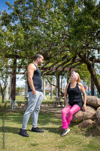 brown athletic boy talking to blonde lady wearing pink pants and black t-shirt, sitting on rock in park on sunny day.
