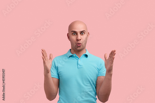 Impressed bald homosexual man with bristle shows big size with both hands, shapes huge object, has eyes wide open, pulls lips, gay friendly, wears blue polo shirt, stands over pink background.