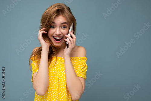 Closeup photo of pretty positive amazed young blonde woman wearing stylish yellow summer dress standing isolated over blue background holding and talking on mobile phone with sincere emotions looking