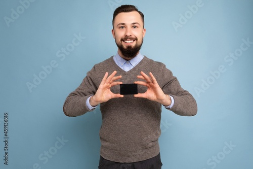 Cool smiling brunette bearded man wearing grey sweater and blue shirt isolated on background wall holding credit card looking at camera photo