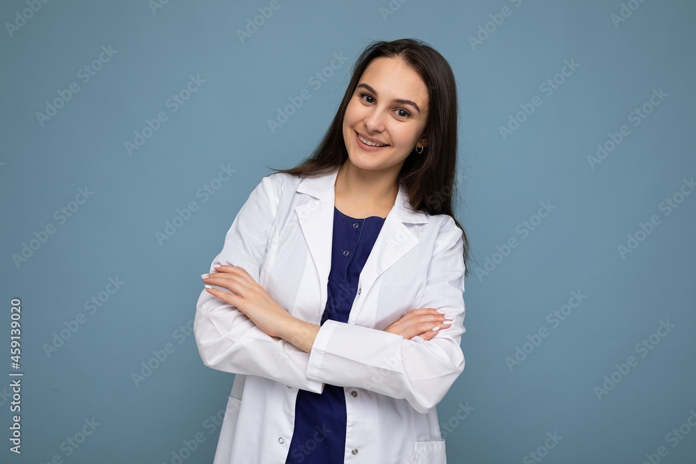 Photo portrait of young attractive beautiful positive smiling brunette woman with sincere emotions wearing white medical coat isolated over blue background with copy space and holding crossed arms Photo portrait of young attractive beautiful positive smiling brunette woman with sincere emotions wearing white medical coat isolated over blue background with copy space and holding crossed arms